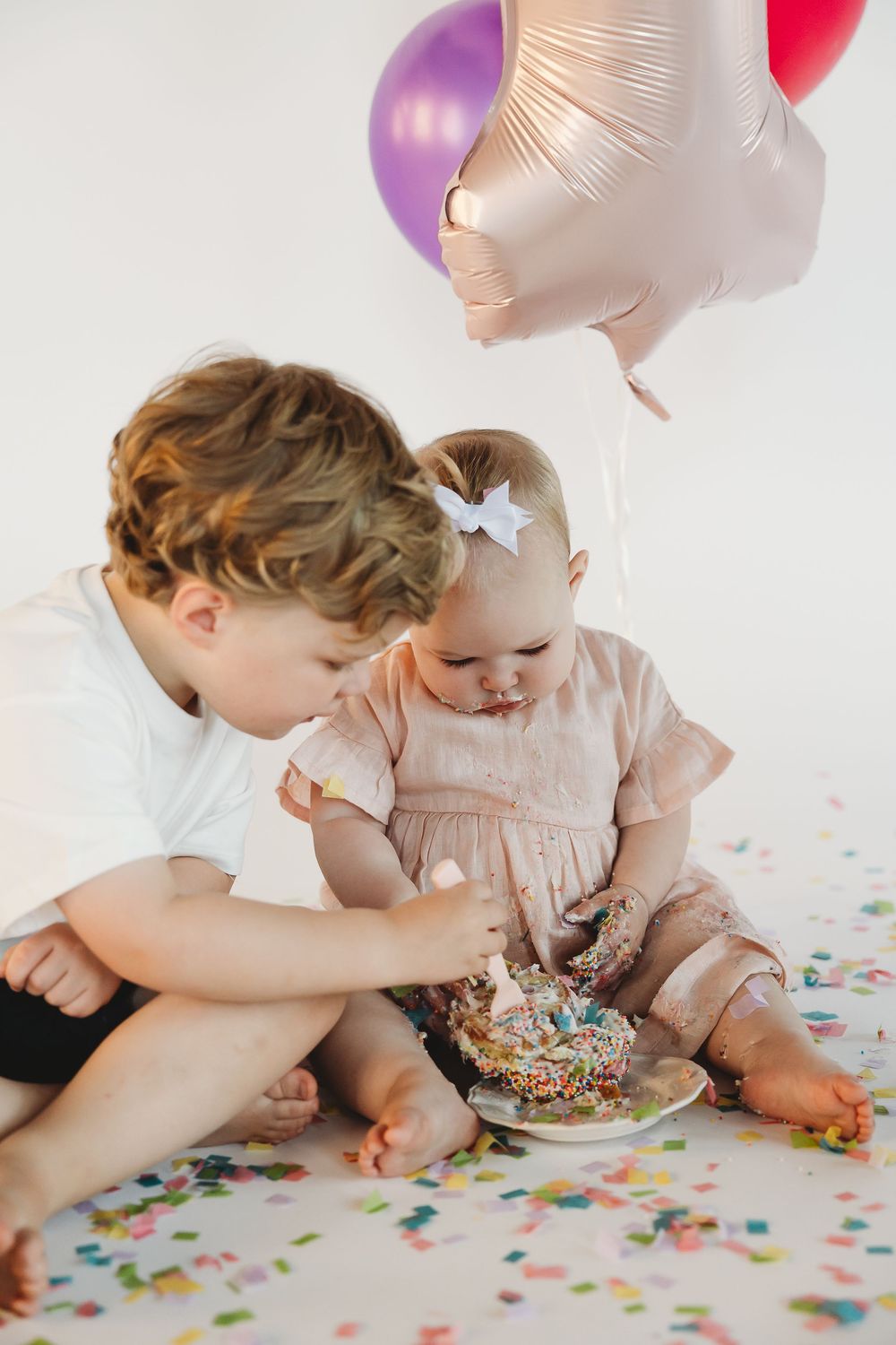 A toddler girl in pink smashes her first birthday cake while her older brother helps, surrounded by colorful balloons.
