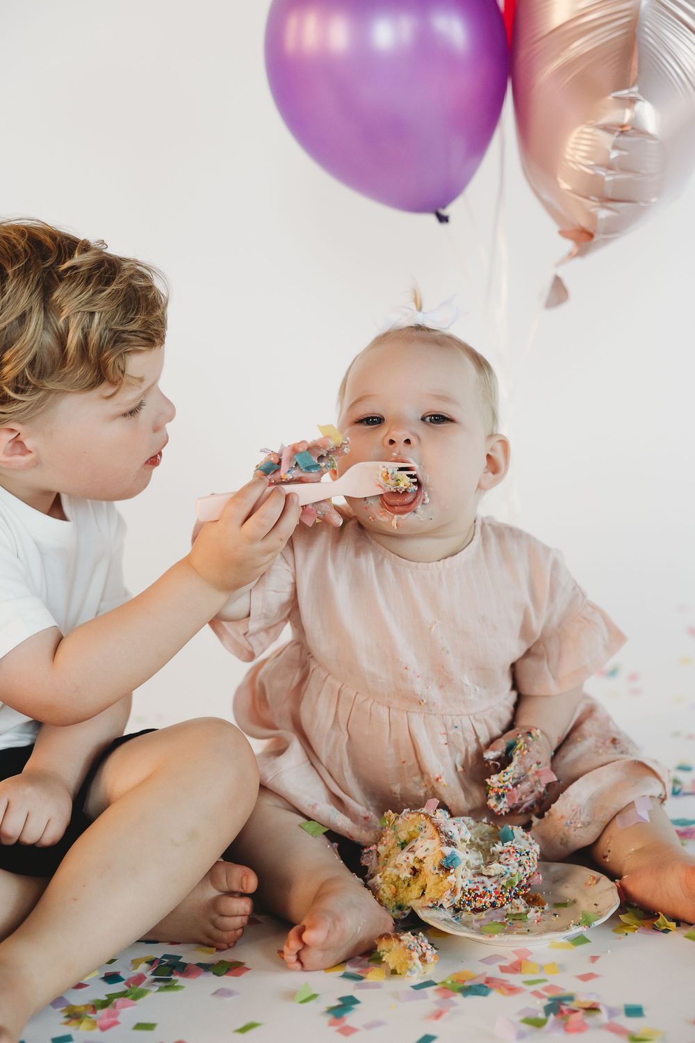Two young children celebrating a first birthday, with older boy feeding baby girl cake surrounded by colorful balloons and confetti.