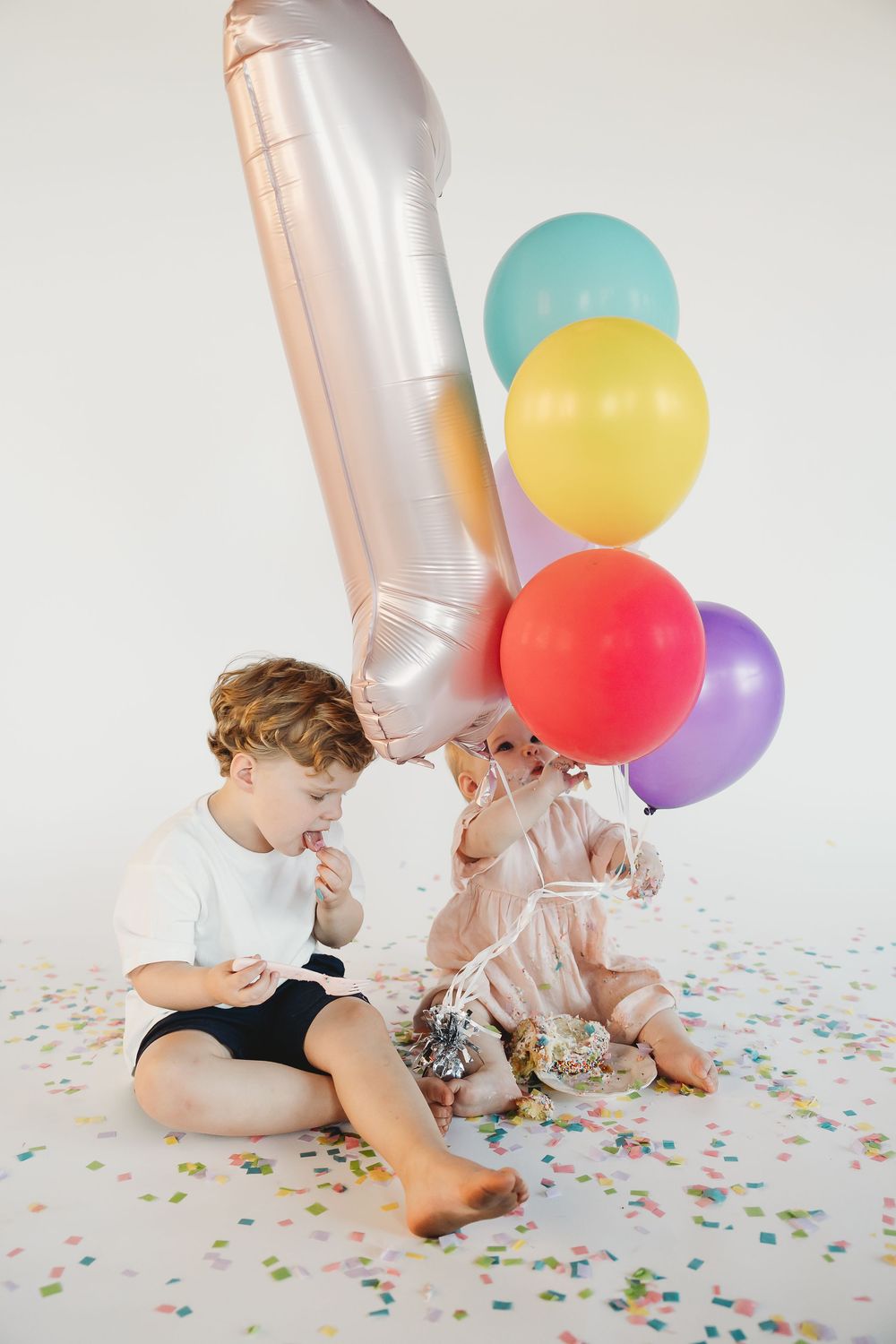 Two young children sitting on floor with colorful balloons and confetti celebrating a first birthday party.