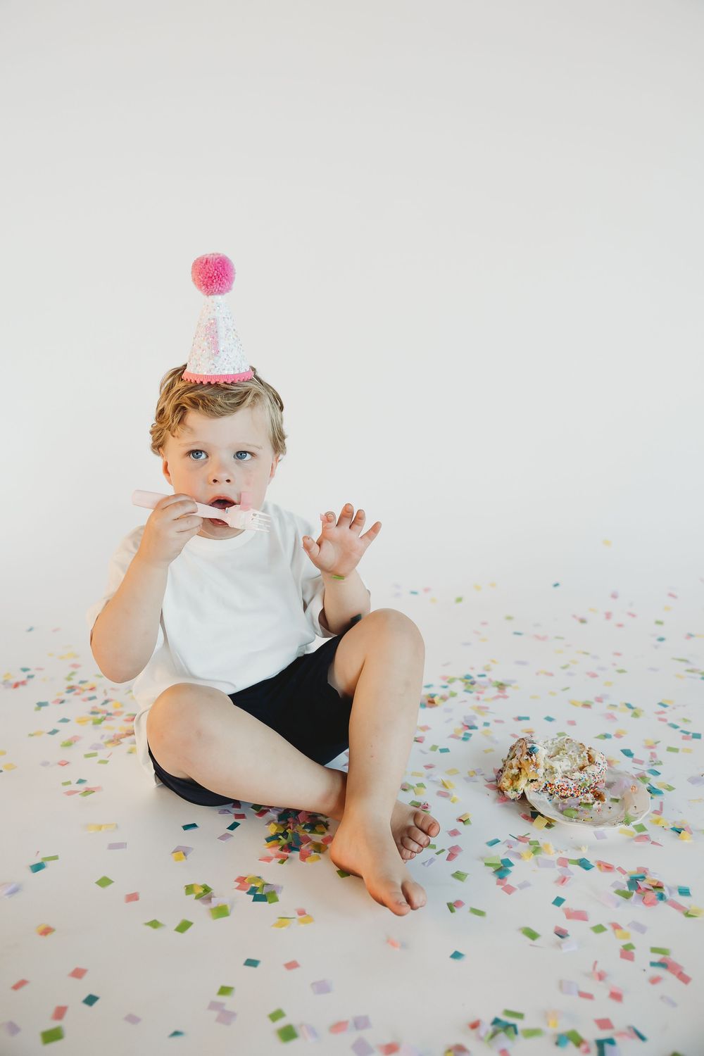 A toddler in a party hat sits on a white floor surrounded by colorful confetti next to a smashed birthday cake.