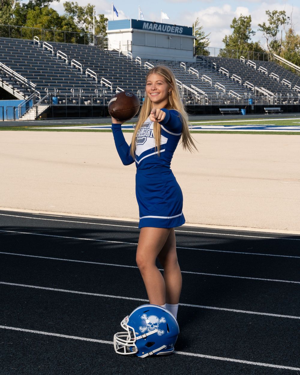 Cheerleader in blue uniform holding football on track field with Marauders stadium bleachers in background.