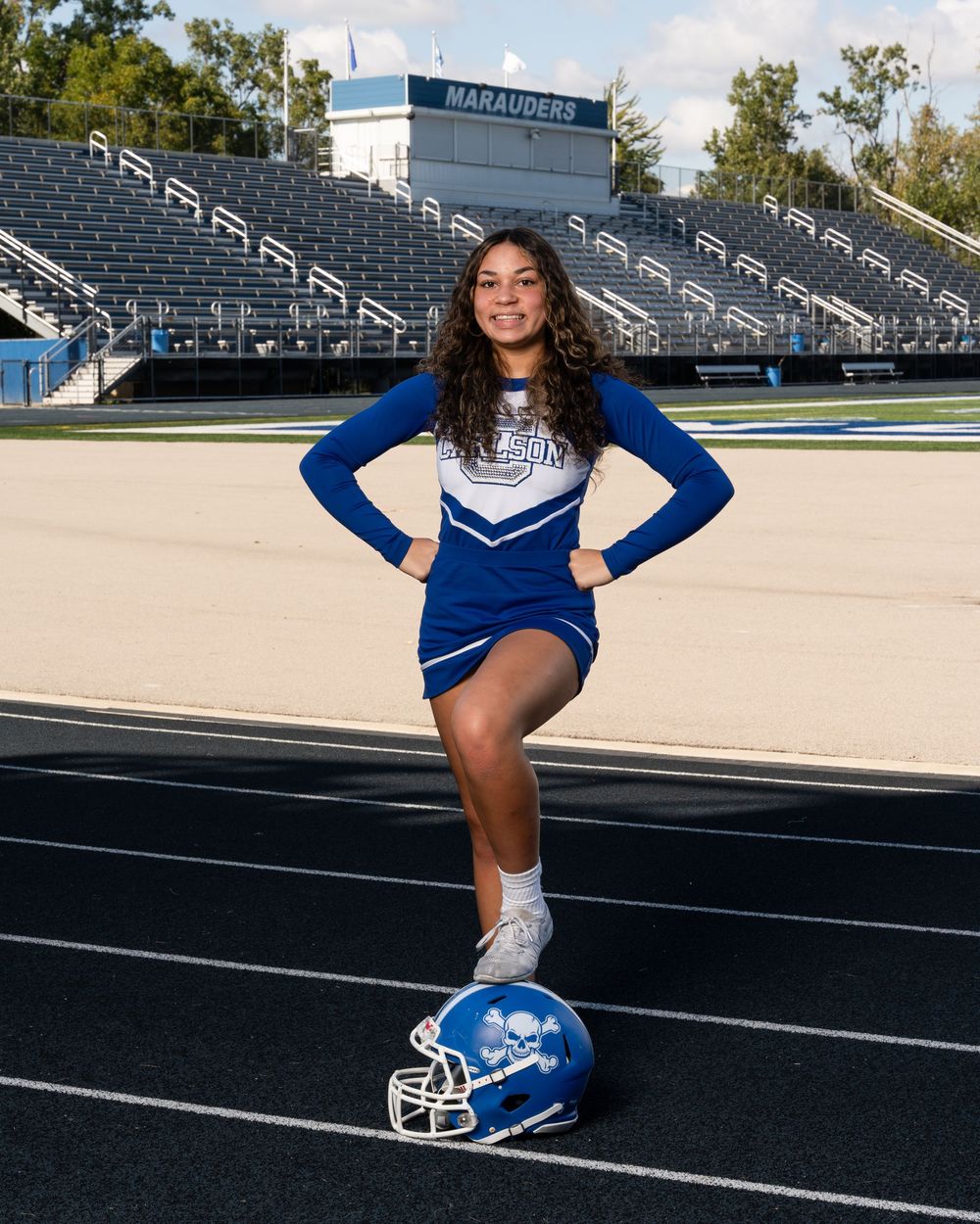 Cheerleader in blue uniform poses on football track with helmet at Marauders stadium.