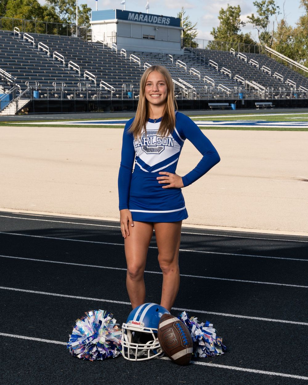Cheerleader in blue uniform poses on football track with helmet and pom-poms, Marauders stadium in background.