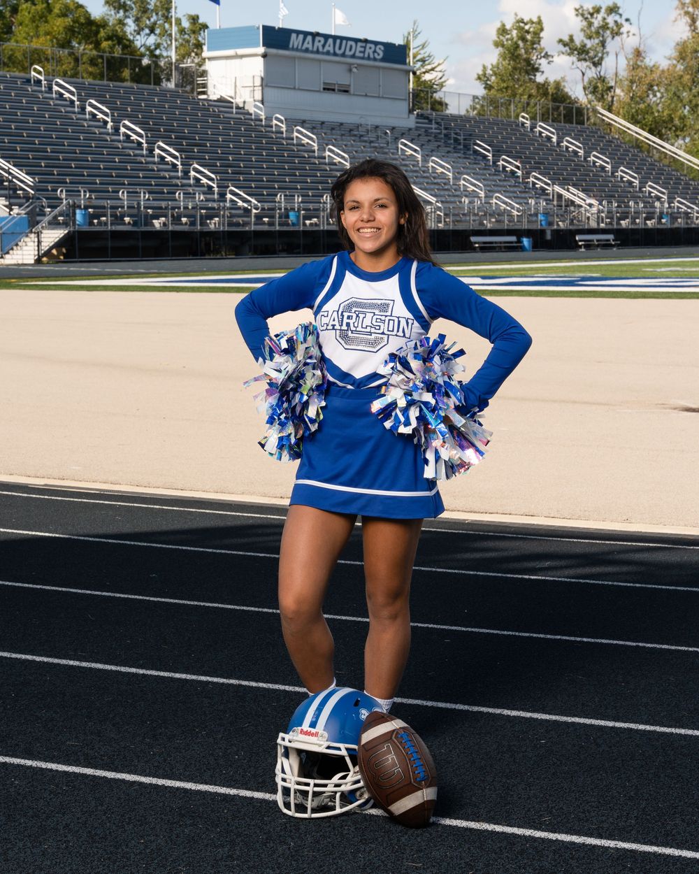 Cheerleader in blue uniform poses on football track with helmet and football, stadium bleachers visible behind her.