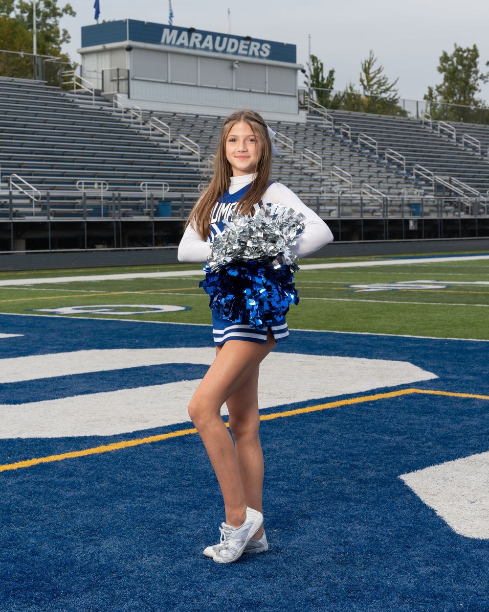 Young cheerleader in blue and white uniform holding pom-poms on a football field with Marauders stadium in background.