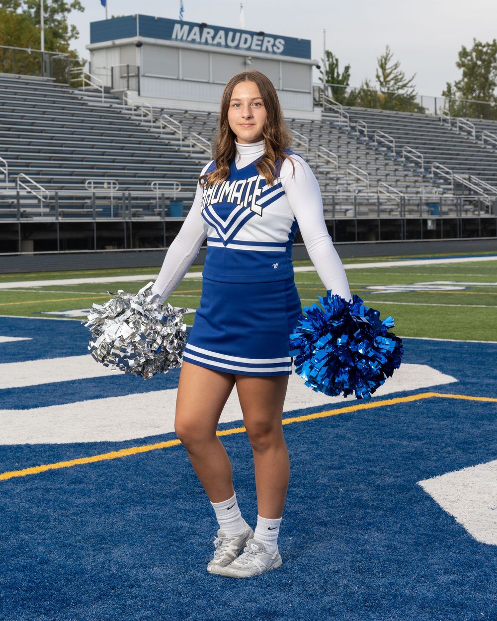 Cheerleader in blue and white uniform holding pom-poms on a football field in front of Marauders stadium.