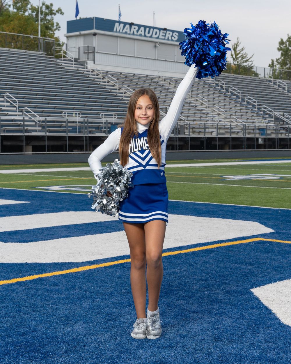 Young cheerleader in blue and white uniform holds pom-poms on a football field in front of Marauders stadium.