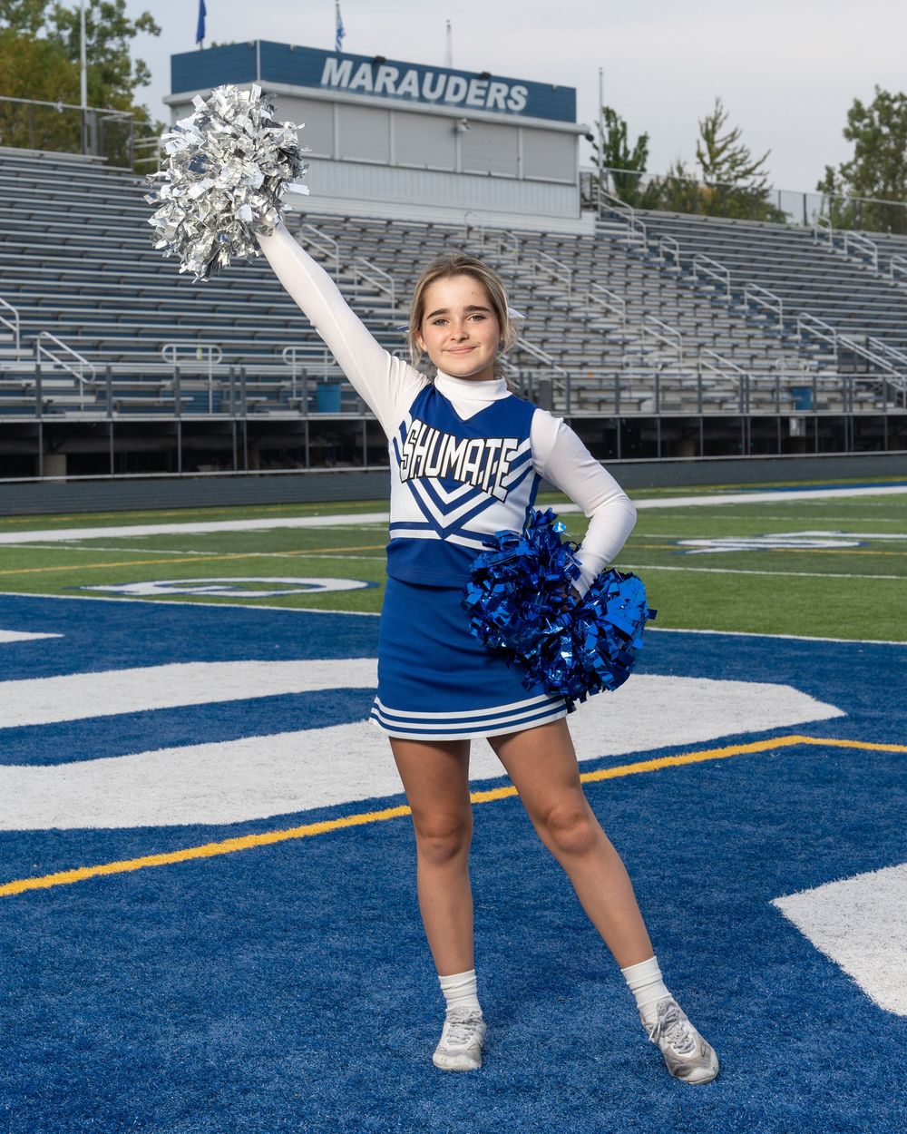 A cheerleader in blue and white uniform poses with silver and blue pom-poms on a football field.