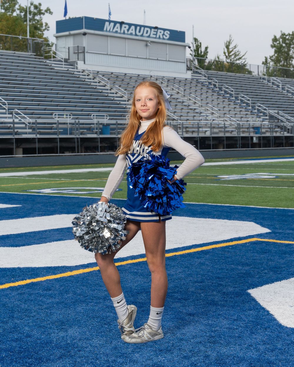 Young cheerleader in blue uniform with pom-poms poses on football field in front of Marauders stadium bleachers.
