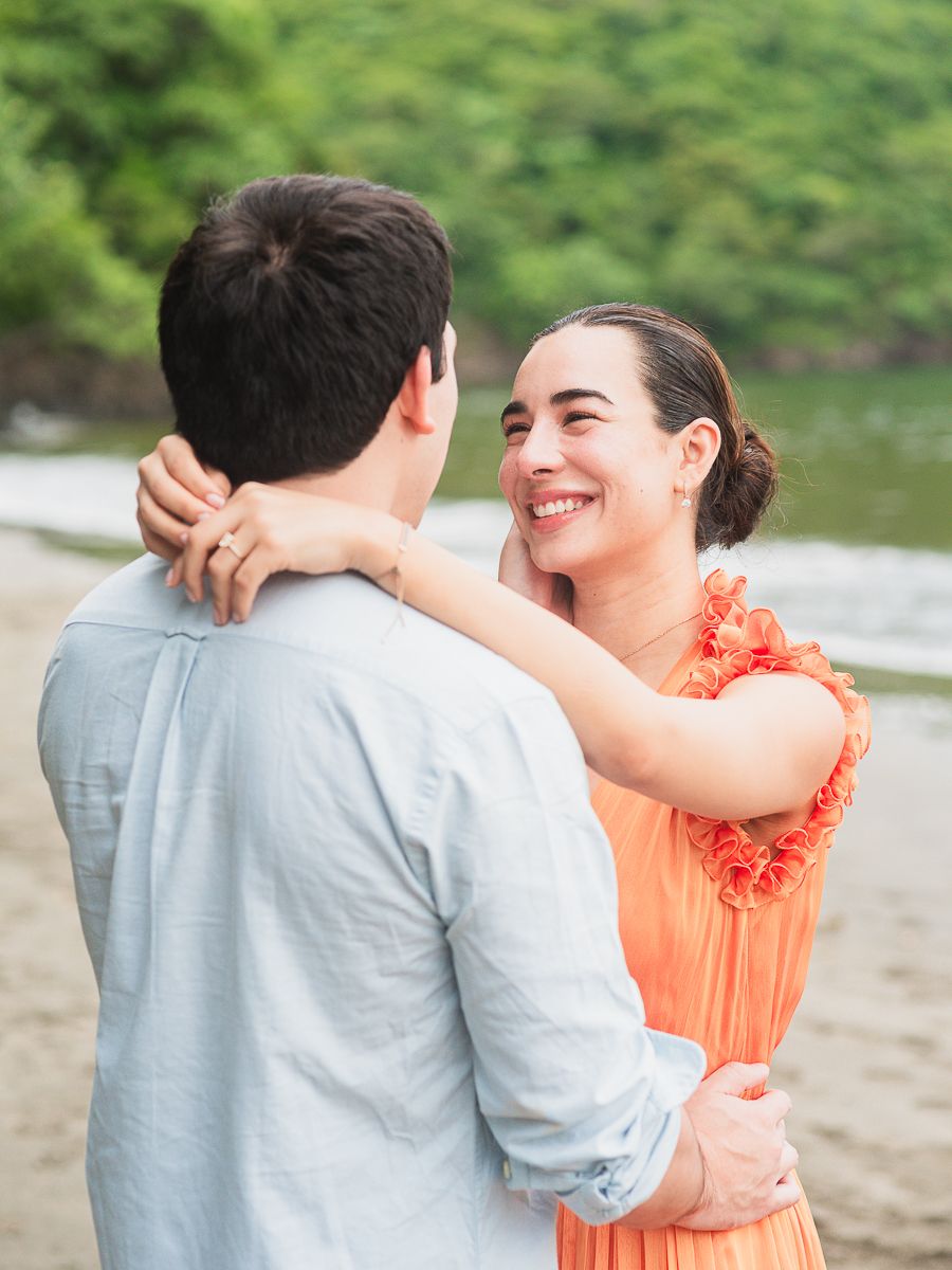 A smiling woman in an orange dress embraces a man on a beach with lush green trees in the background.