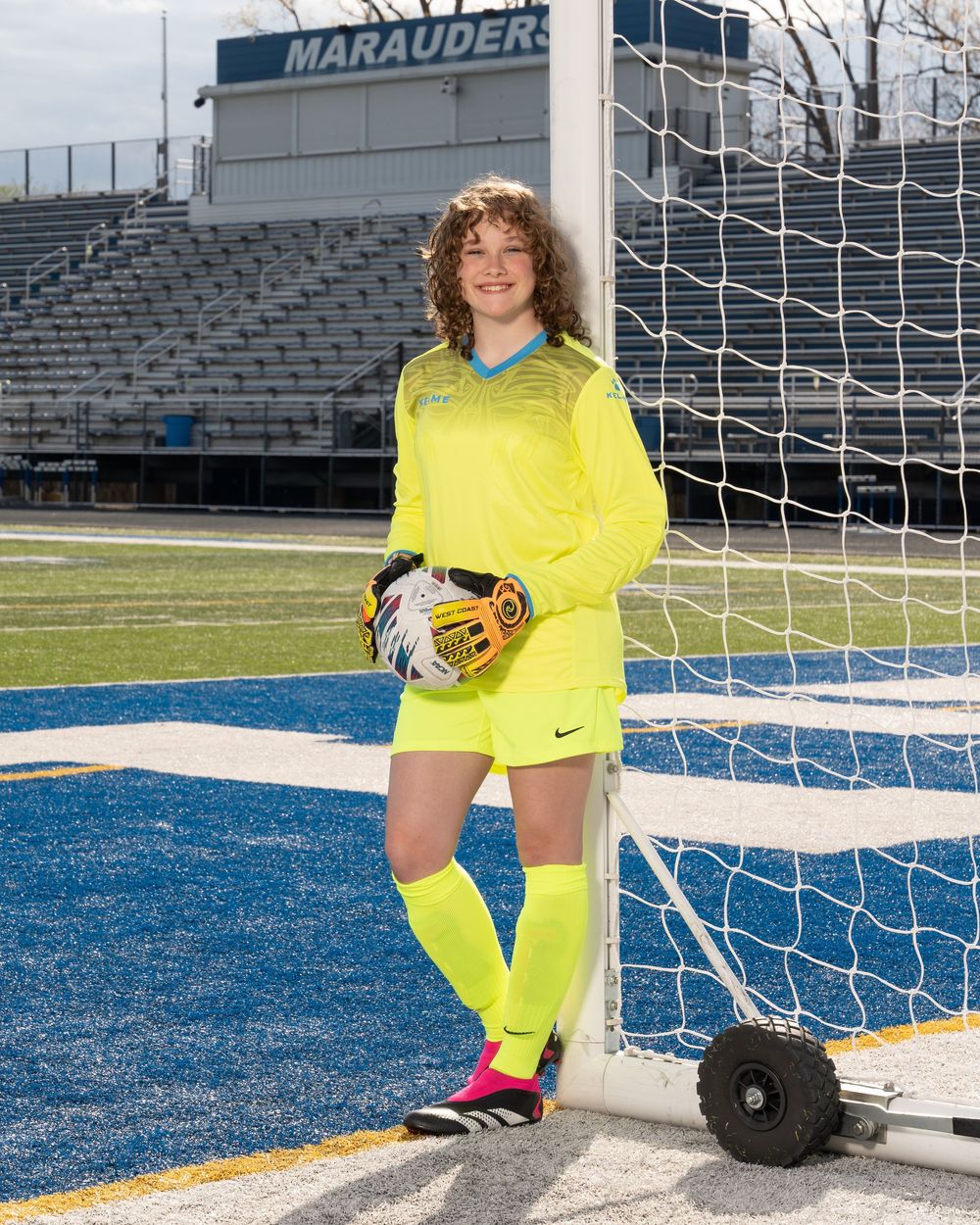 Female soccer goalkeeper in neon yellow uniform poses by goal net on blue turf field at Marauders stadium.