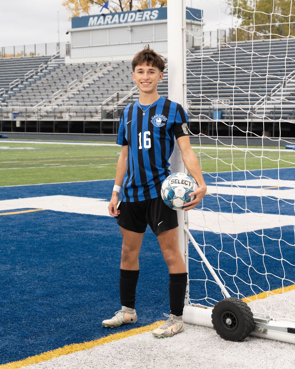 Soccer player #16 in blue and black striped jersey holds a Select ball near goal net on turf field with Marauders stadium.