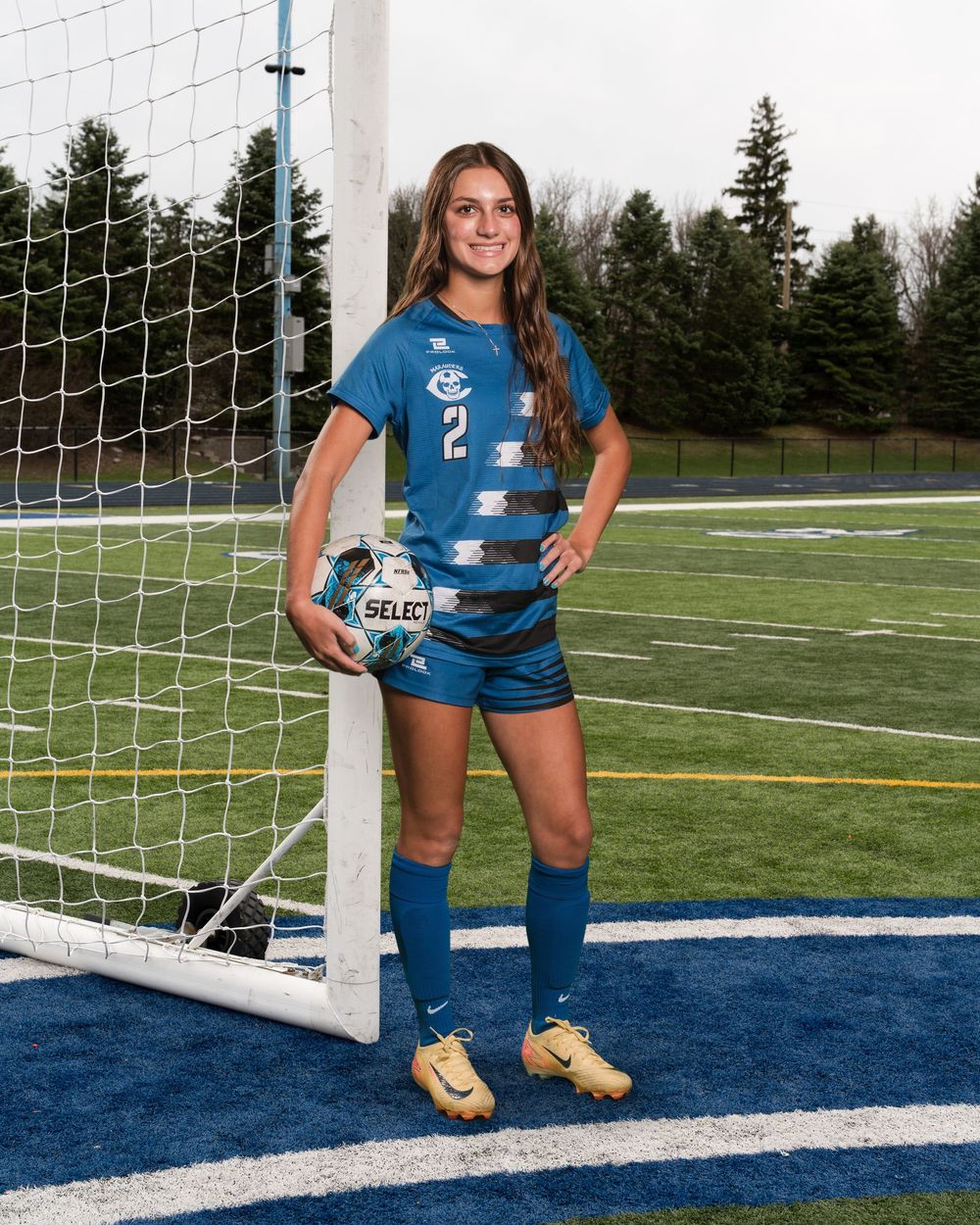 Female soccer player #2 in blue uniform holds a Select ball while posing by a goal post on a green field.