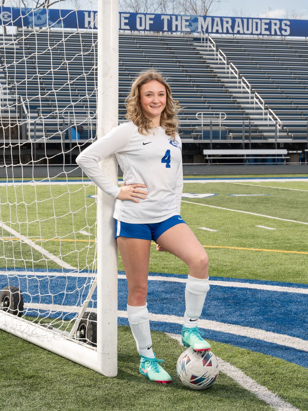 Female soccer player #4 posing by goal post on turf field, wearing white Nike jersey and blue shorts.