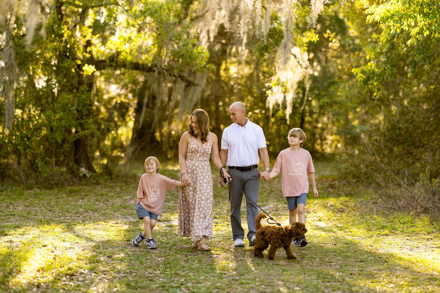 Happy family of four holding hands and walking a dog through a sunlit southern forest path.