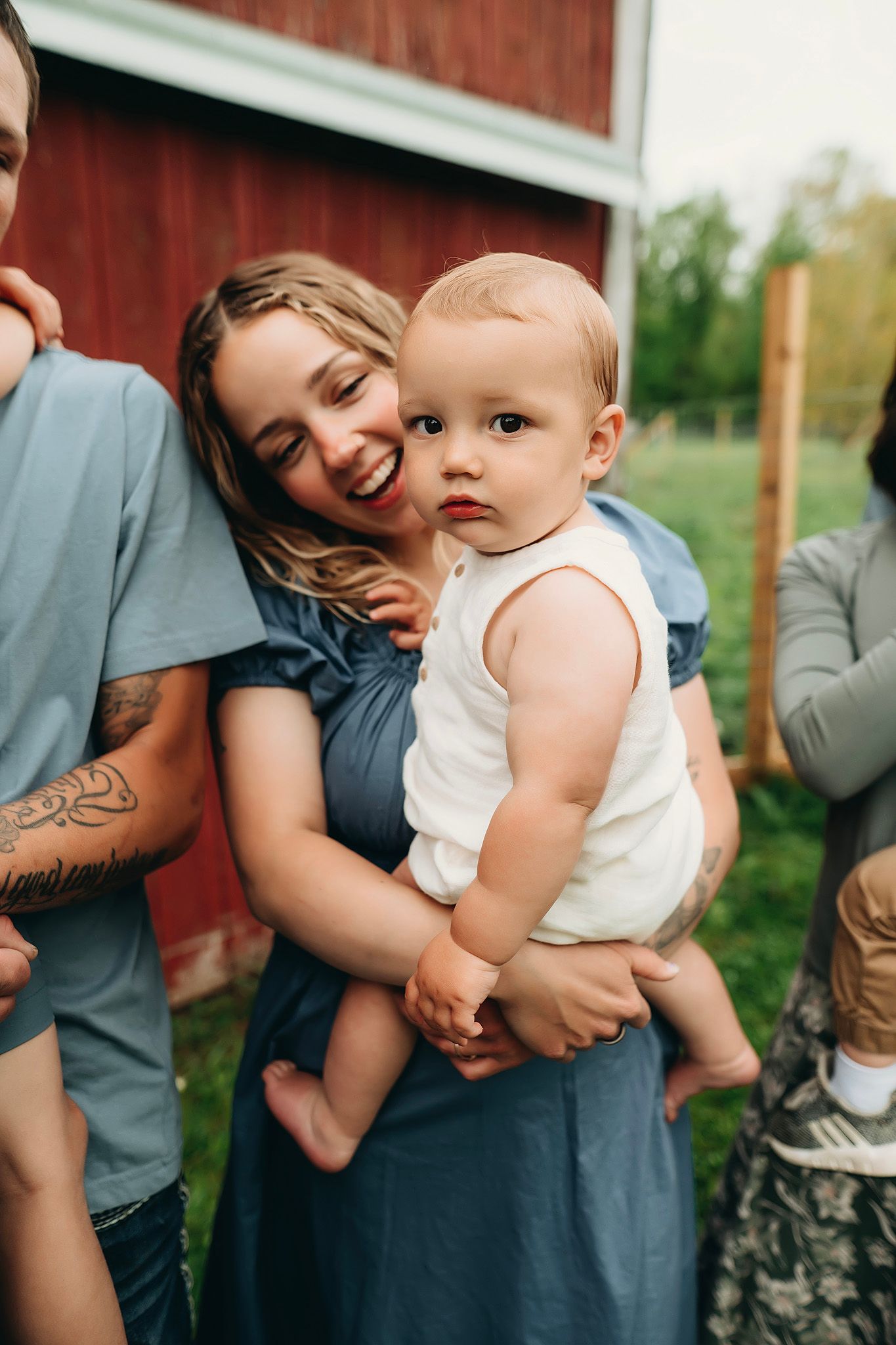 A smiling woman holds a curious baby in a white onesie outdoors near a red barn on a green farm.