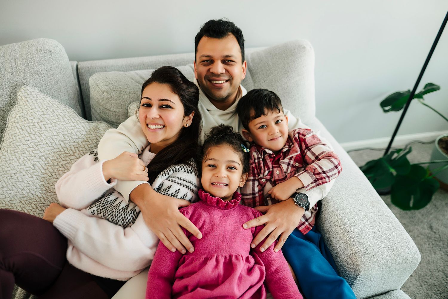 Happy family of four smiling together on a cozy gray sofa at home, parents hugging two young children.
