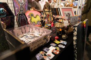 Colorful art vendor booth displaying handmade crafts, jewelry, and pop culture artwork at an indoor convention market.