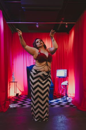 A plus-size woman in a red bra and chevron skirt poses with arms raised in a red-curtained room.