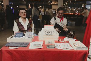 Two vendors in vintage attire sit at a red-draped table displaying prints and merchandise at an indoor market event.
