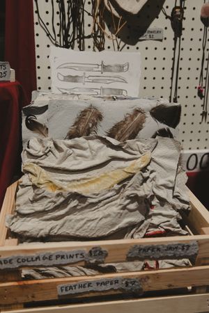Handmade feather paper and collage prints displayed in a wooden crate at an artisan market stall.