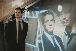 A young man in a black suit stands beside a large poster of suited men at an event.