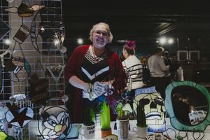 A smiling woman with glasses arranges colorful artwork at an indoor vendor booth.