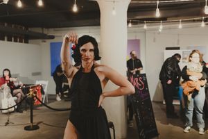 Performer in black dress posing confidently at an indoor art event with band and guests in background.
