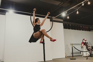 Female aerial performer in black dress and red heels spinning on a lyra hoop in a white gallery space.