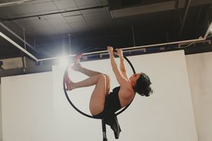 Aerial acrobat in black outfit performing inverted pose on a lyra hoop suspended from ceiling in studio.