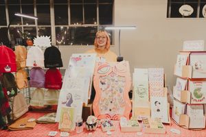 A smiling woman stands behind a craft fair table displaying colorful illustrated cards and art prints at an indoor market.