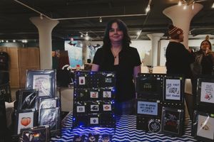 A vendor stands behind a display table with art prints and illuminated merchandise at an indoor market event.