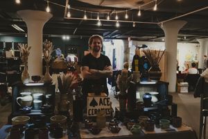 A craftsman stands proudly behind a display of handmade ceramics at ACE Ceramics booth in a dimly lit artisan market.
