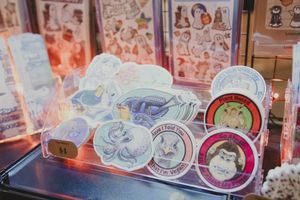 Colorful stickers and circular pins displayed in a clear acrylic stand at a vendor table.