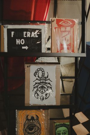 Art prints on metal shelf including scorpion drawing and funeral home sign artwork.