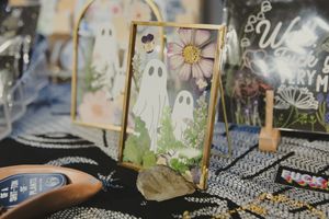 A framed ghost and floral illustration displayed on a black table at an art vendor booth.