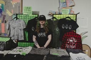 A tattooed woman with green hair stands behind a table of graphic tees at an art market.