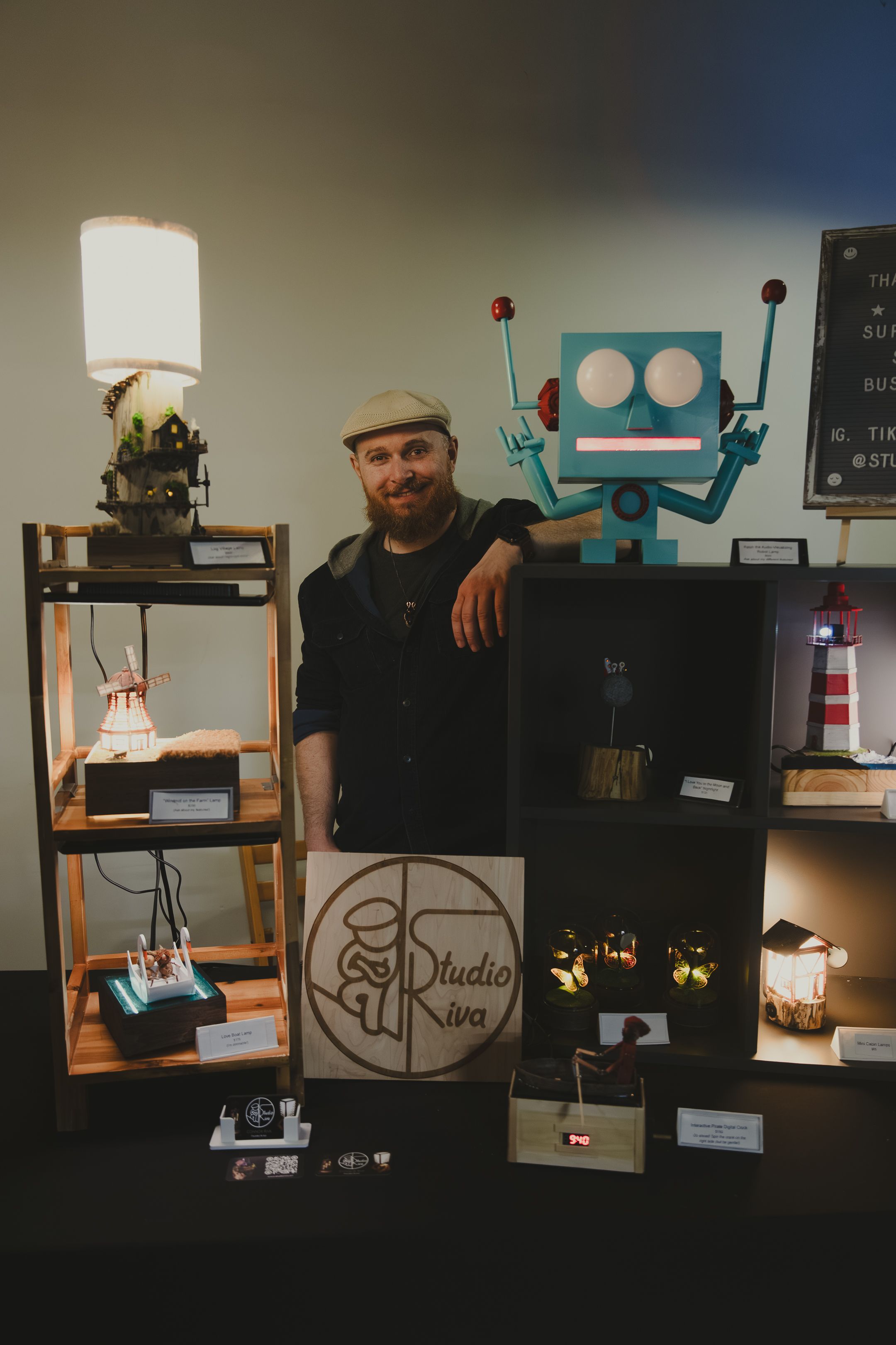 Smiling bearded man at Studio Iva craft booth with handmade lamps and robot art.