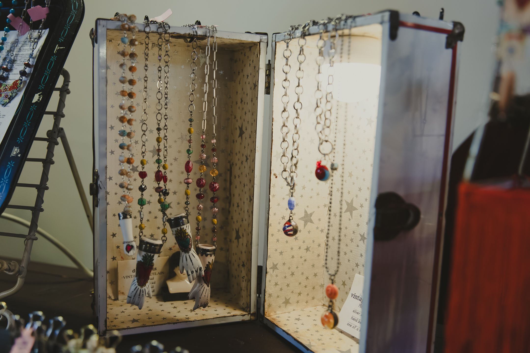 Open jewelry display case showcasing colorful necklaces and earrings hanging neatly inside a vintage suitcase at a craft fair.