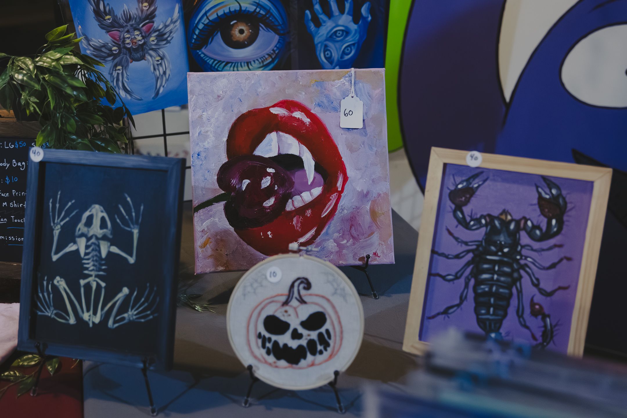 Dark art display featuring skeleton prints, a scorpion painting, lip artwork, and a jack-o-lantern plate at an art market.