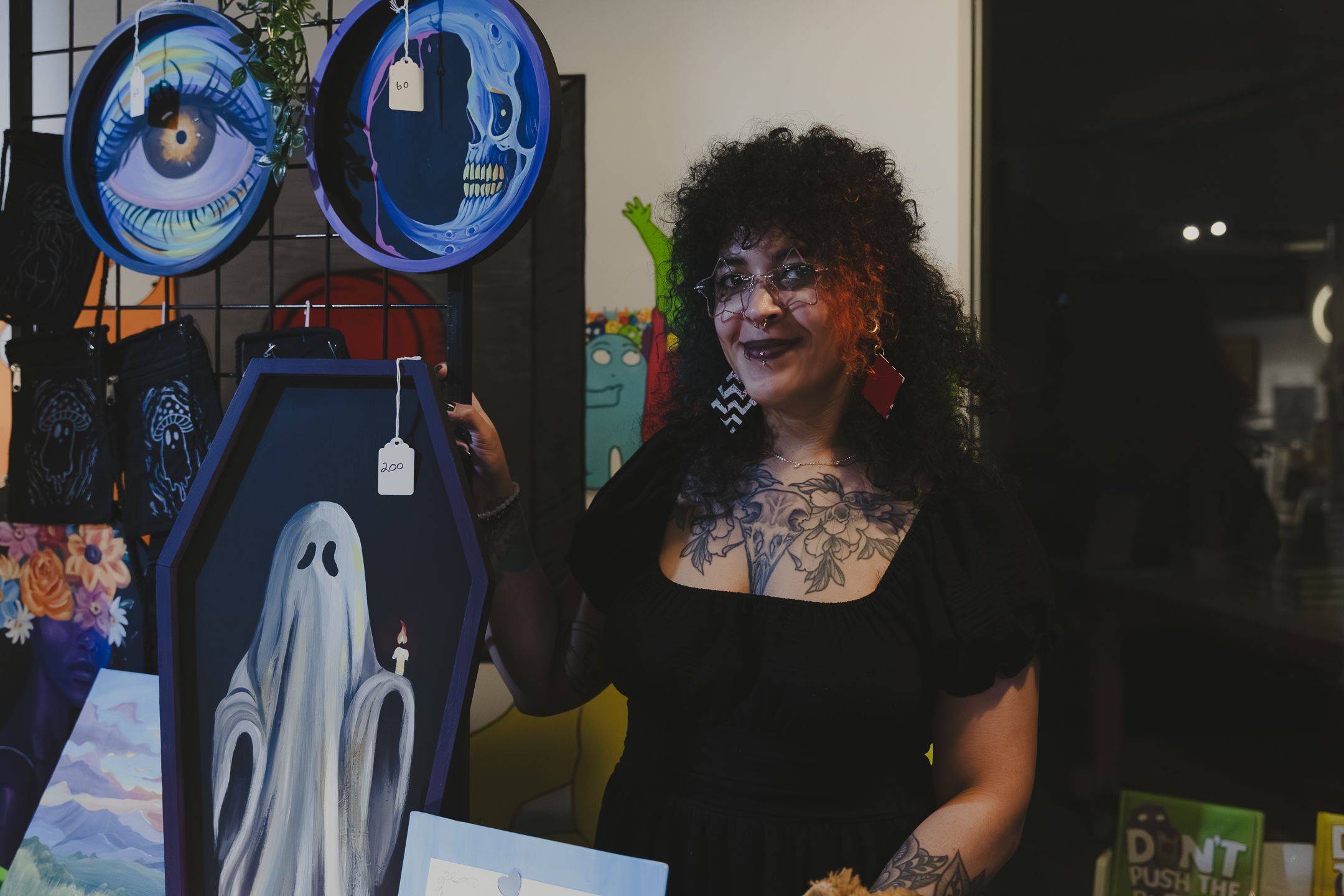 Tattooed woman with curly black hair smiles beside spooky Halloween art including ghost and eye designs at an art fair.