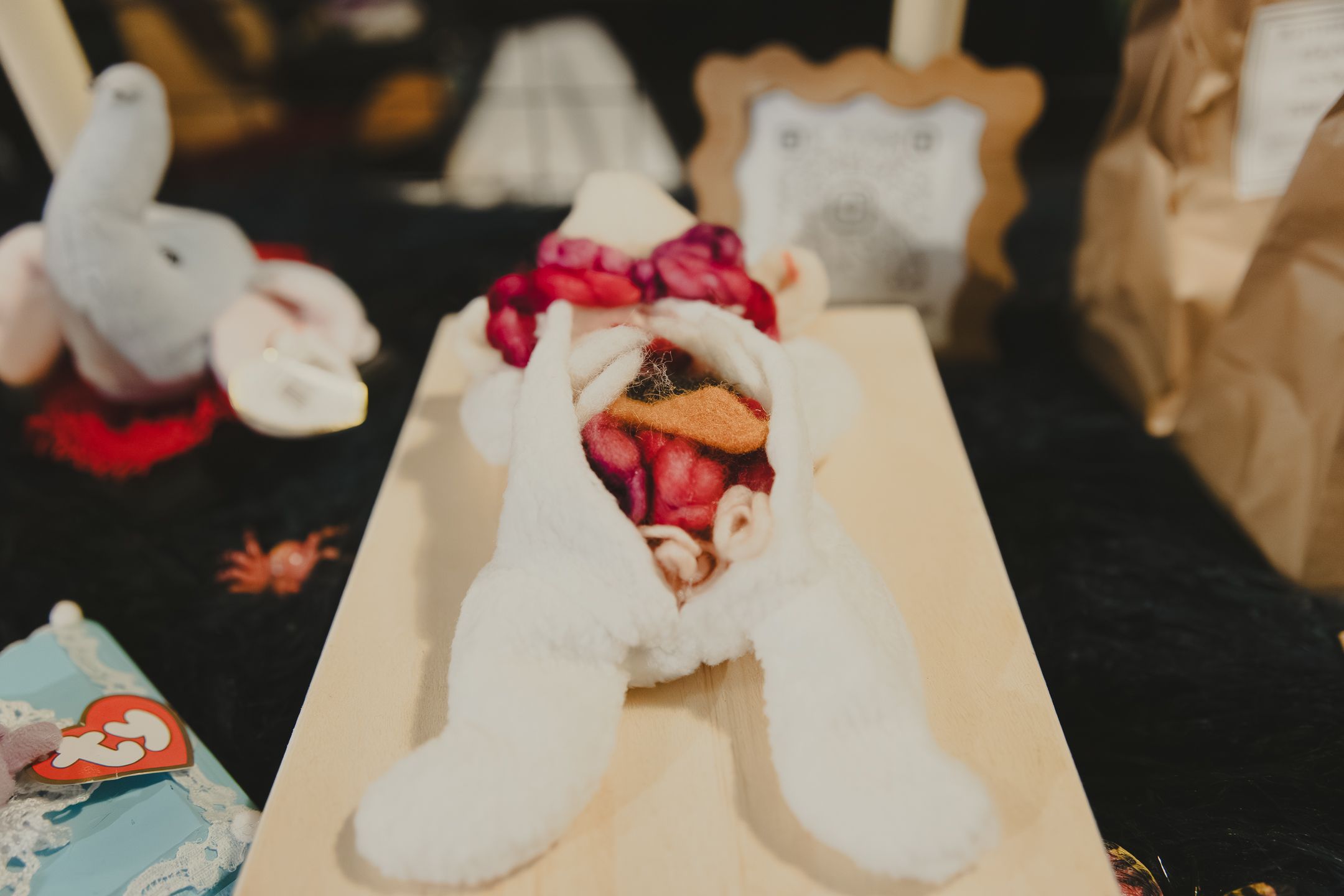White stuffed bunny toy with red bow displayed on wooden tray at craft fair or toy market.