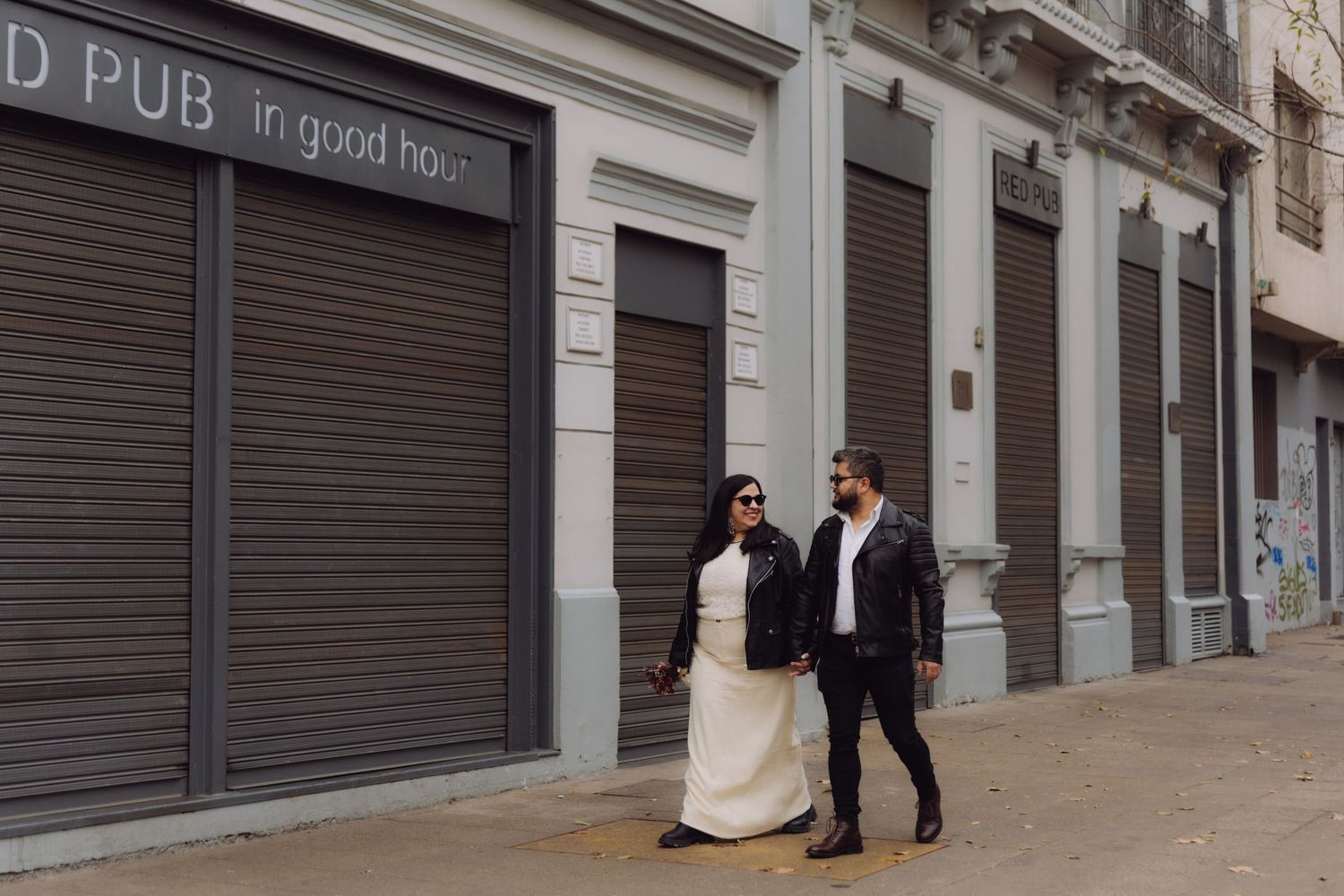 Bride in white dress and groom in leather jacket walk hand-in-hand past Red Pub storefront.