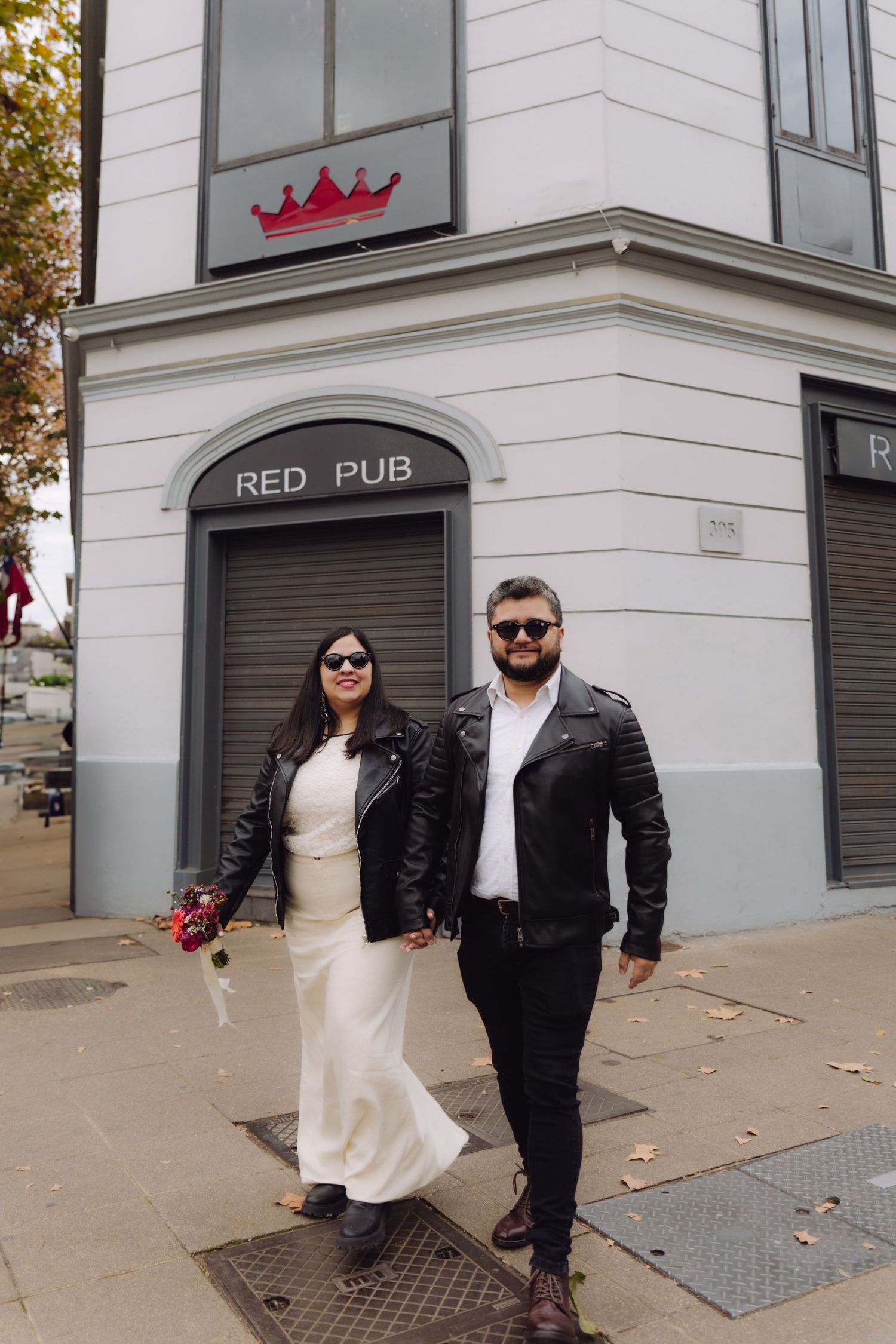 Stylish couple in black leather jackets walk holding hands outside Red Pub on city sidewalk.