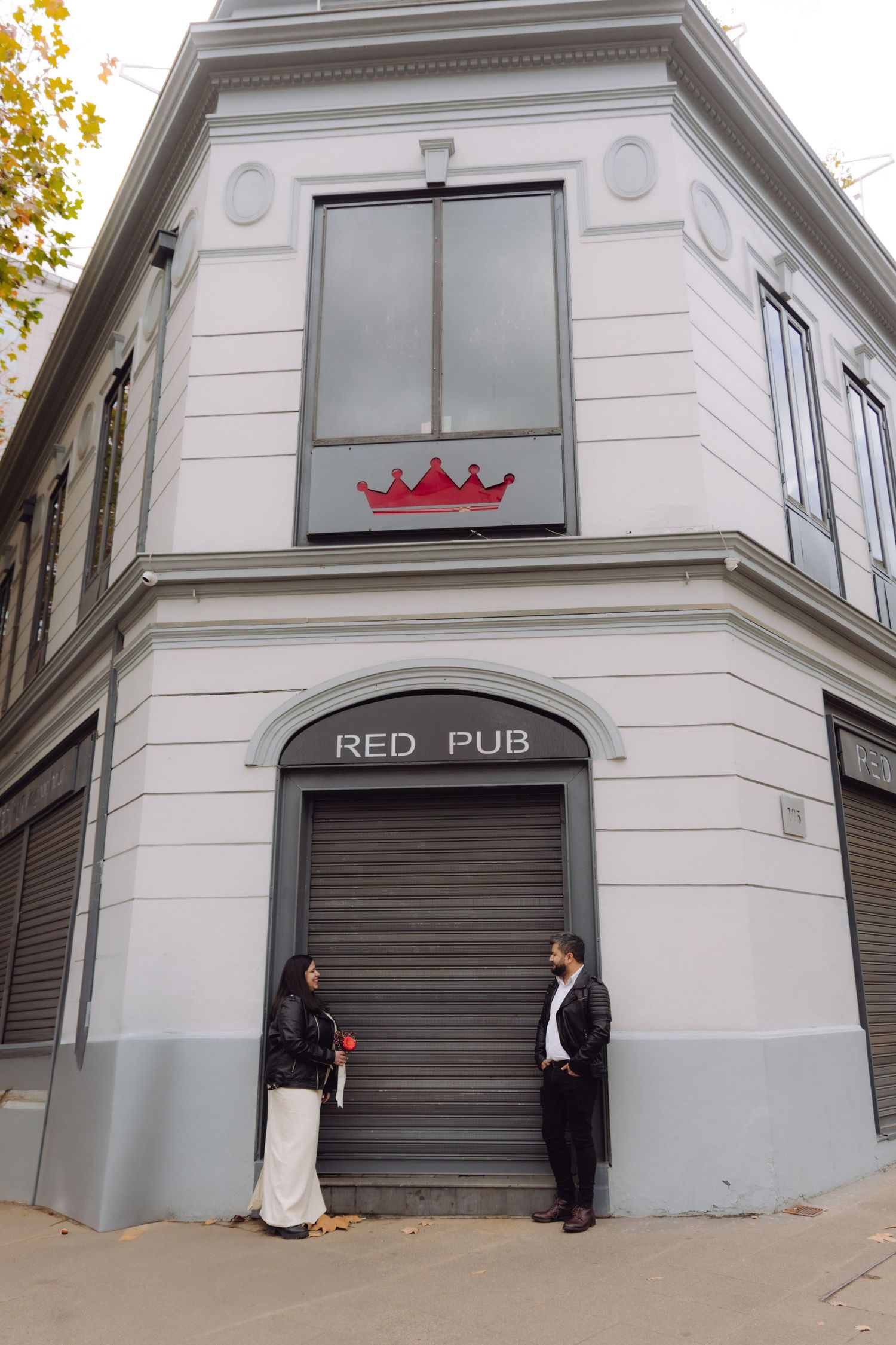 Couple in leather jackets face each other beneath Red Pub sign on elegant white corner building.