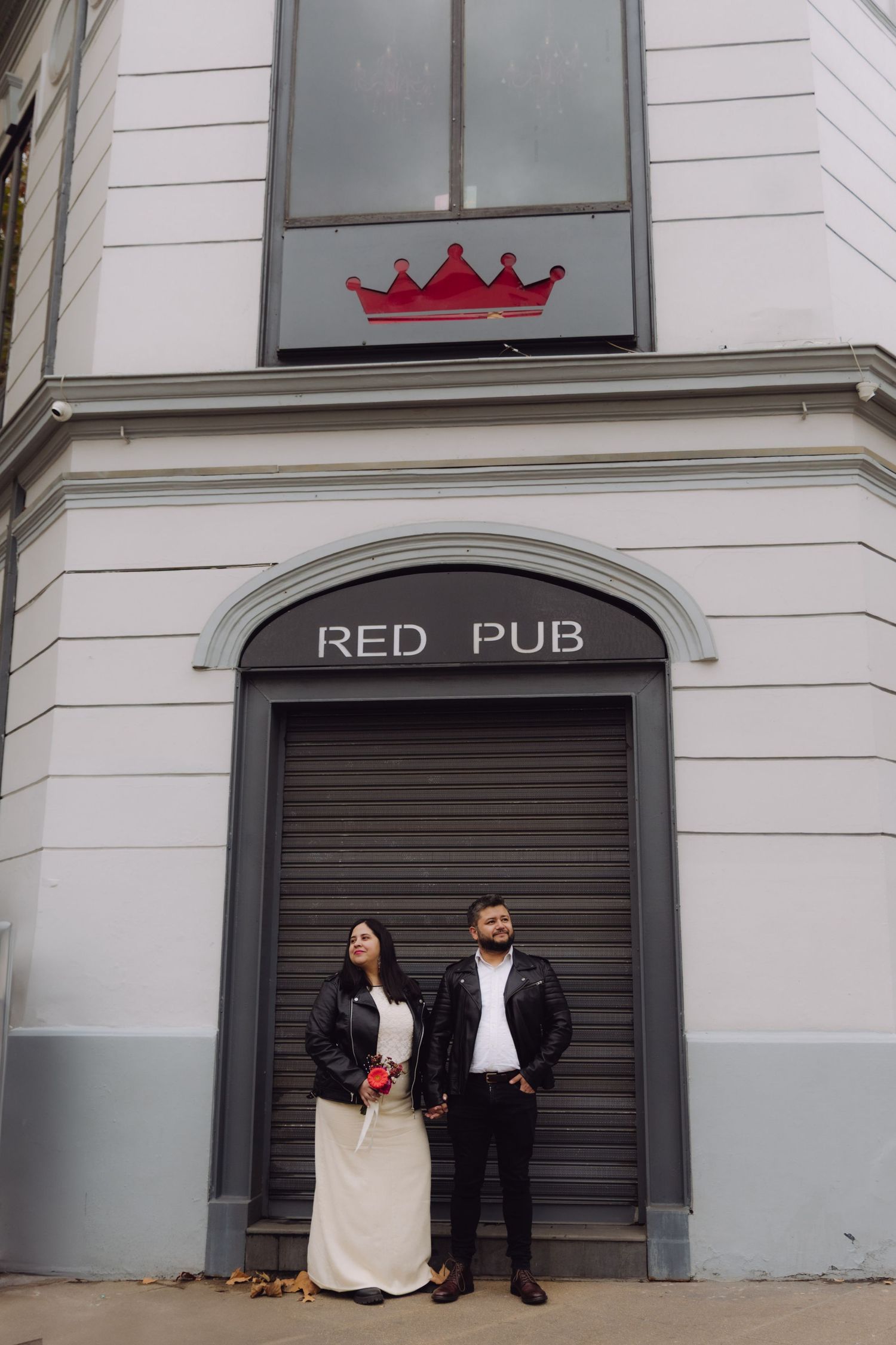 Bride and groom in matching leather jackets stand beneath crown logo at Red Pub building.