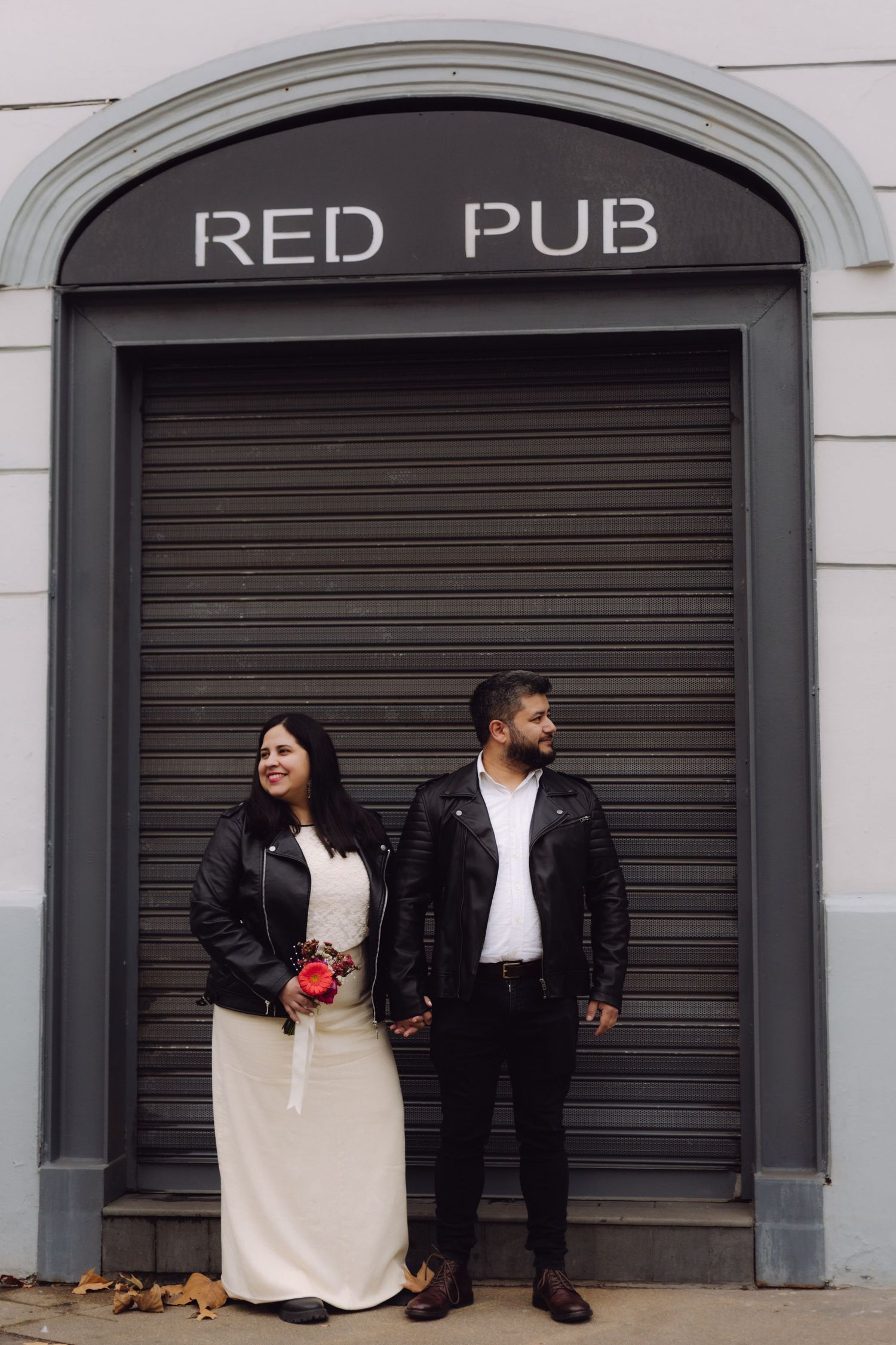 Couple in black leather jackets pose holding hands under arched Red Pub sign on gray wall.