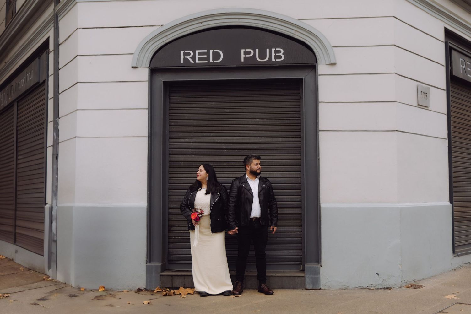 Bride holding red bouquet and groom in leather jackets stand before Red Pub corner building.