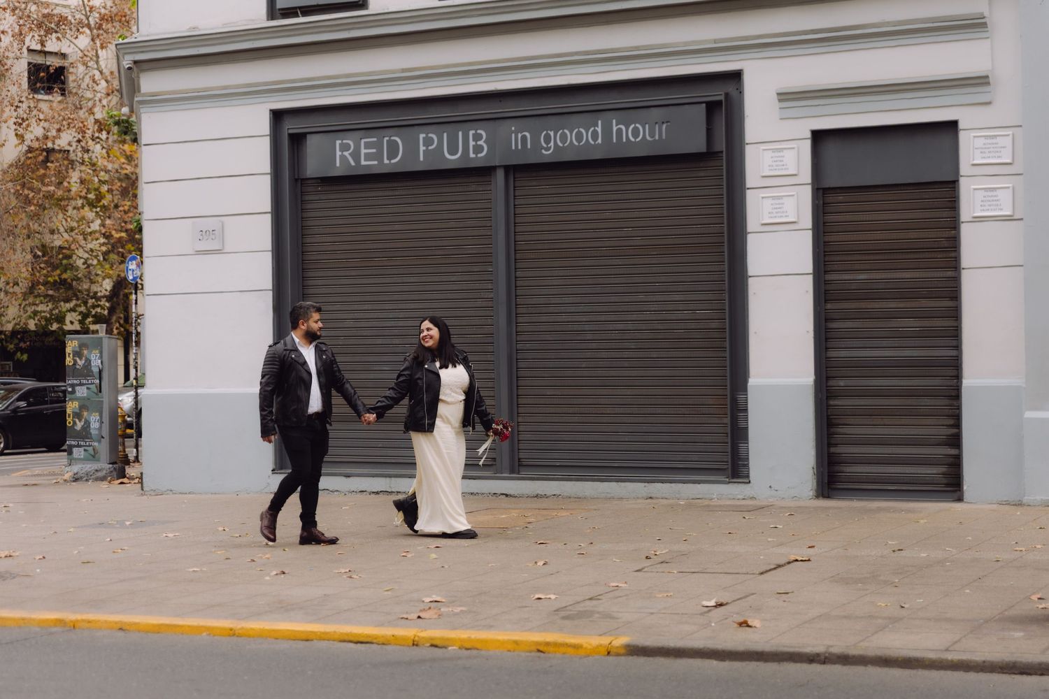 Couple in leather jackets walk hand-in-hand past Red Pub storefront on sunny city street.