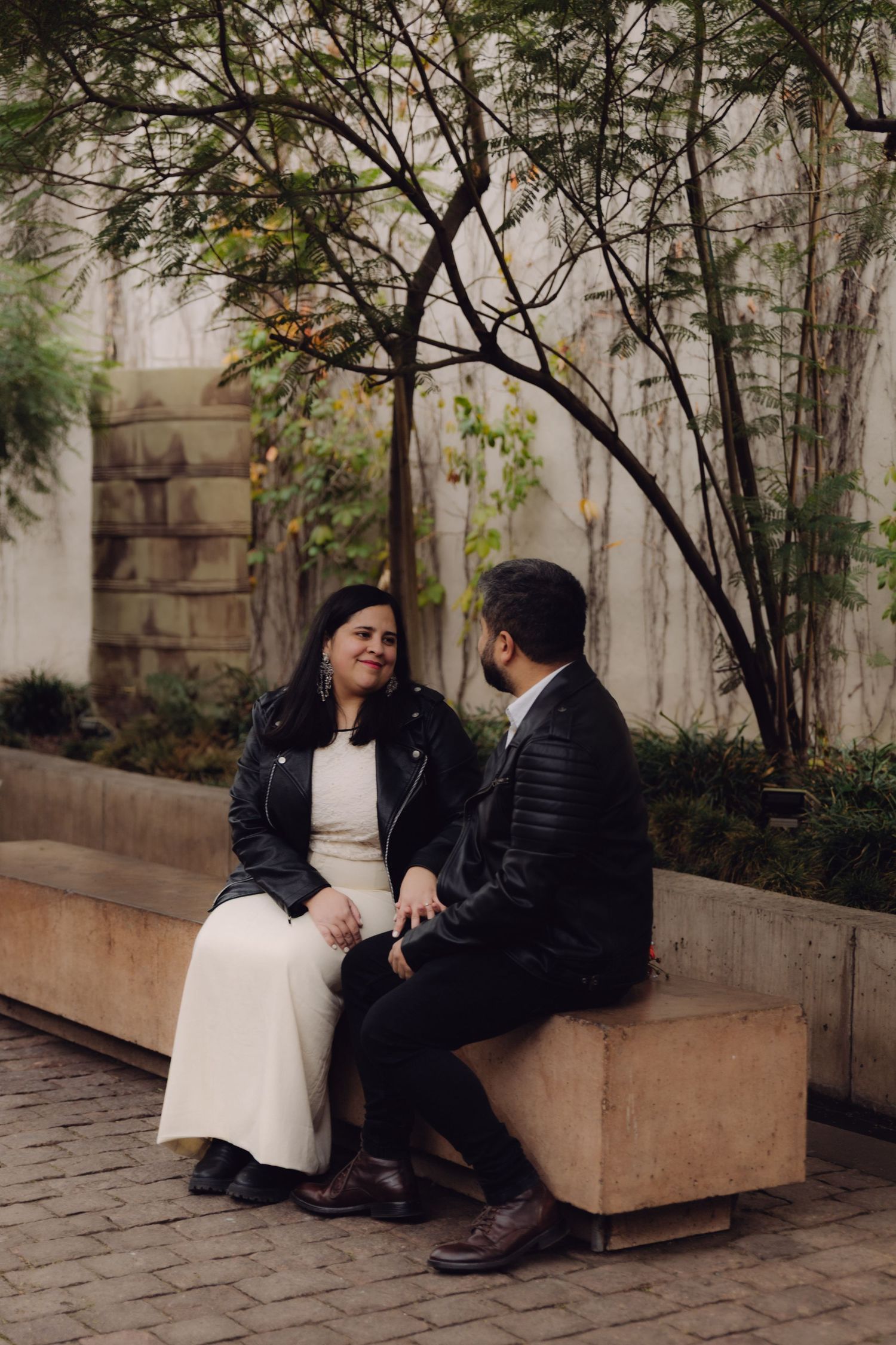 Couple in black leather jackets sit together on outdoor bench surrounded by autumn trees.